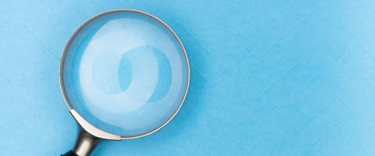 Silver-rimmed magnifying glass with black handle laid upon solid pale blue backdrop.